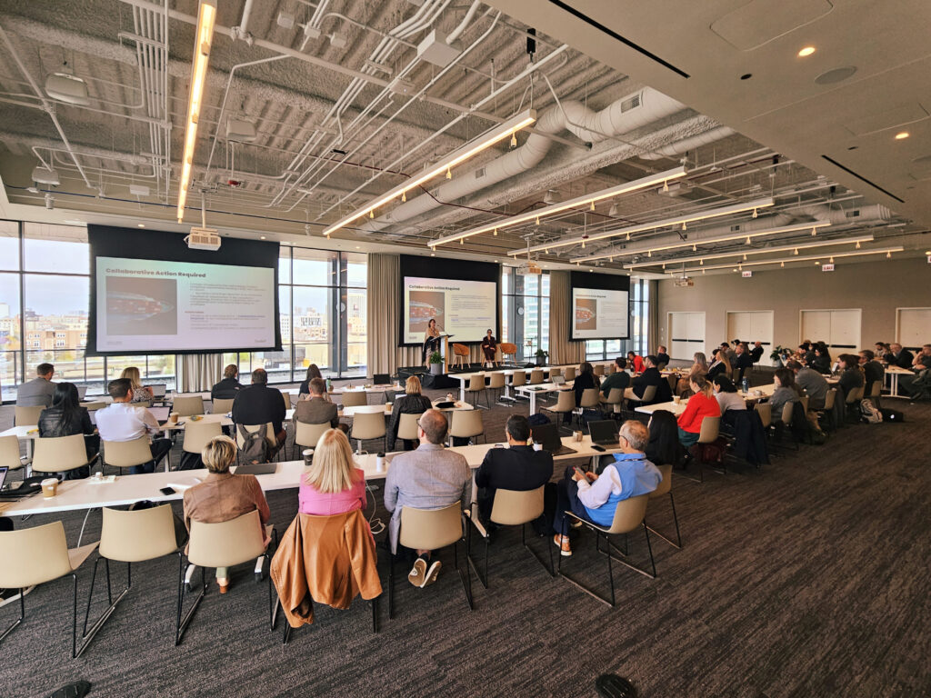 A wide conference room with people in chairs facing a stage and screens.