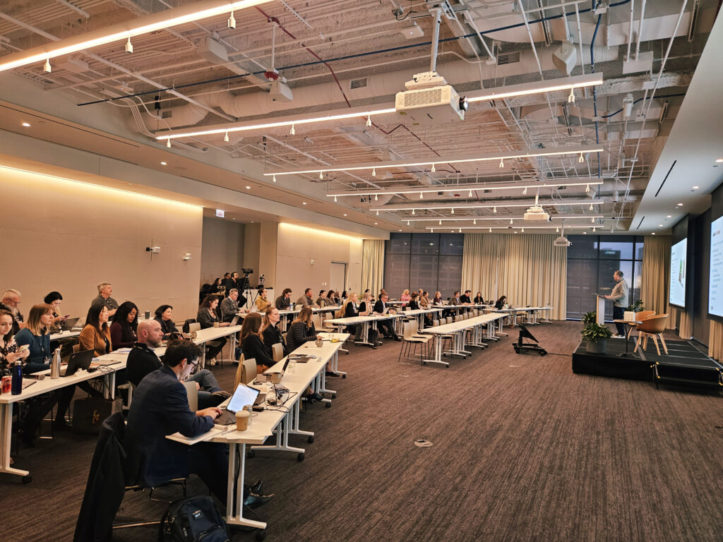 A wide conference room with rows of seated people facing a stage