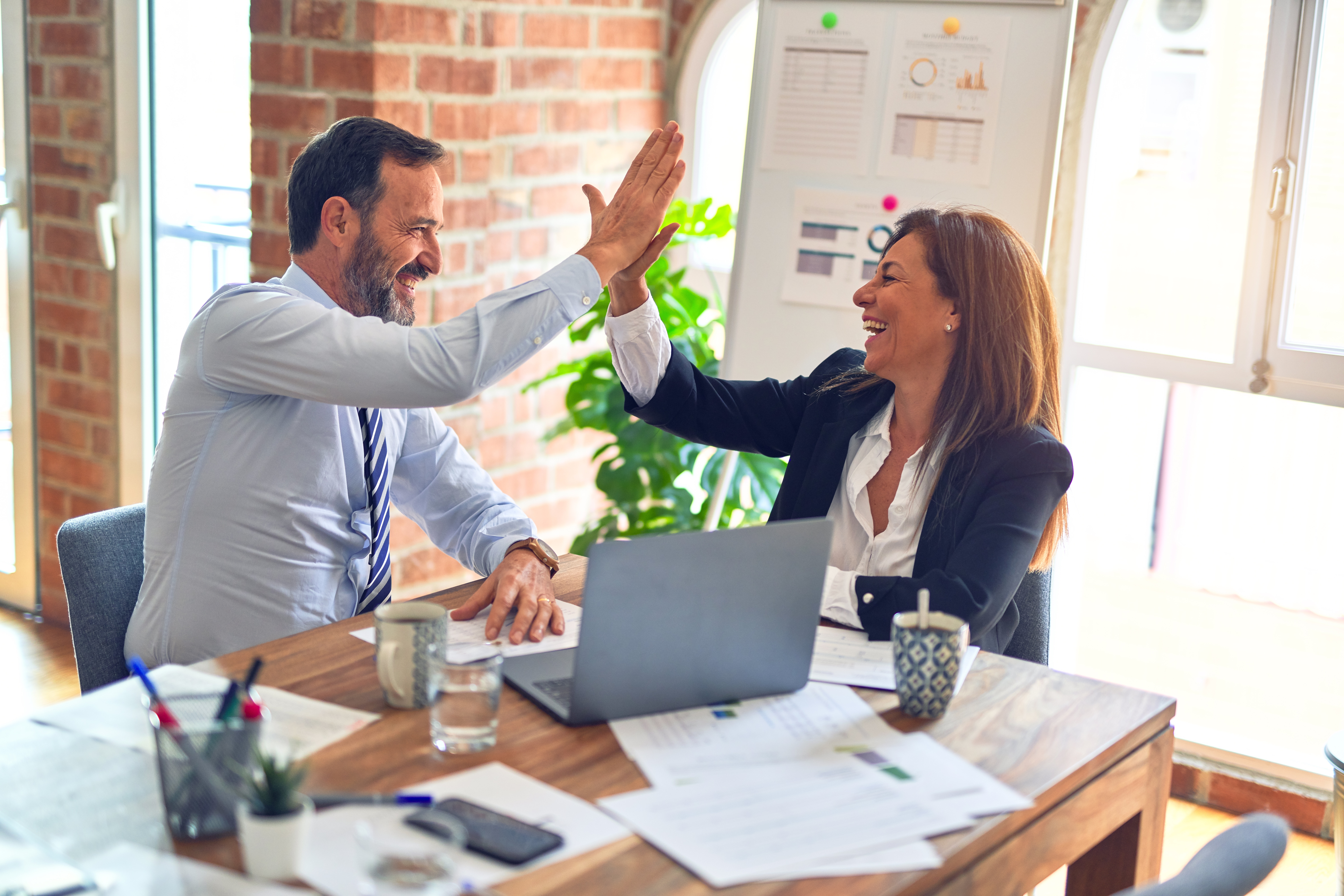 Two colleagues high-fiving at a desk with a laptop and printed reports, celebrating progress on supplier ESG performance